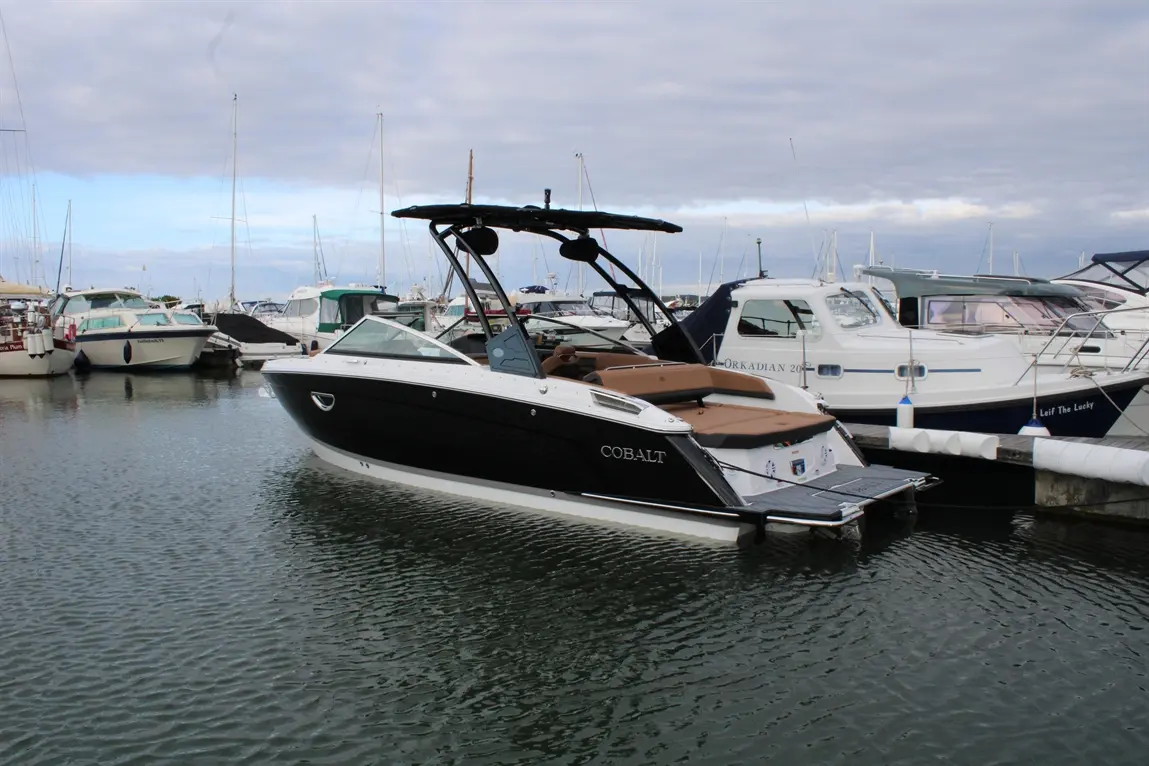 A Cobalt boat docked in a marina, surrounded by other boats, with a cloudy sky reflecting on the water.